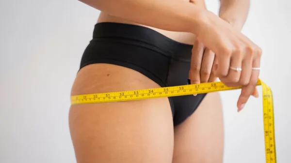A woman in black bikini bottom measuring her waist.